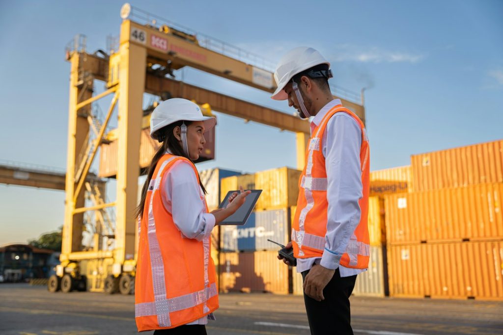 Two logistics workers wearing safety helmets discuss plans near shipping containers